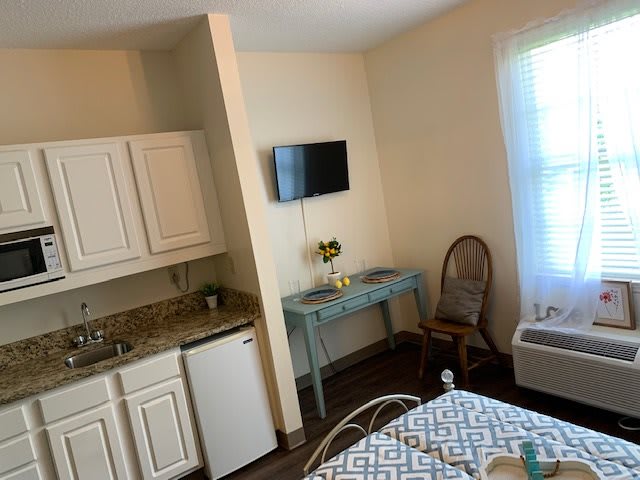 A cozy room with a kitchenette, featuring a bed, wall-mounted TV, desk, chair, and a window.