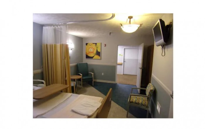 A hospital room with a bed, chairs, a wall-mounted television, and artwork on the wall.