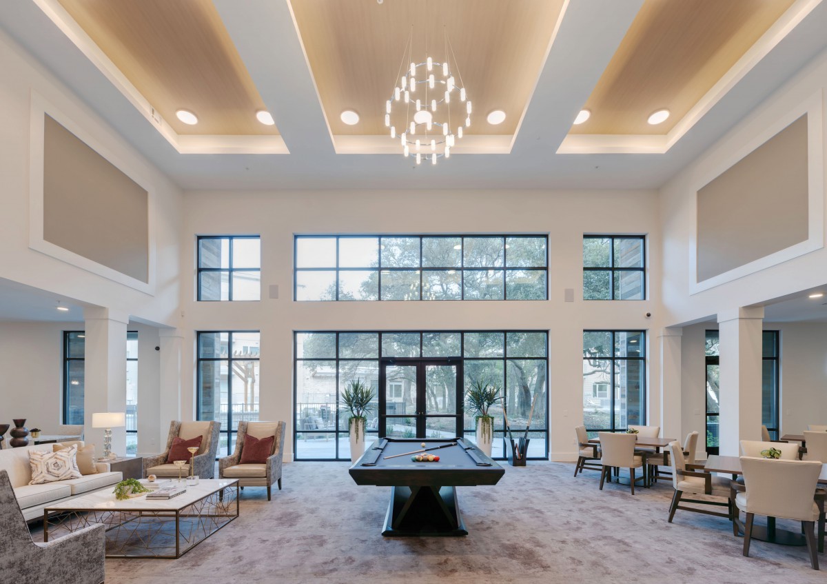 A modern lobby with a billiards table, seating areas, and a large chandelier, illuminated by natural light from expansive windows.