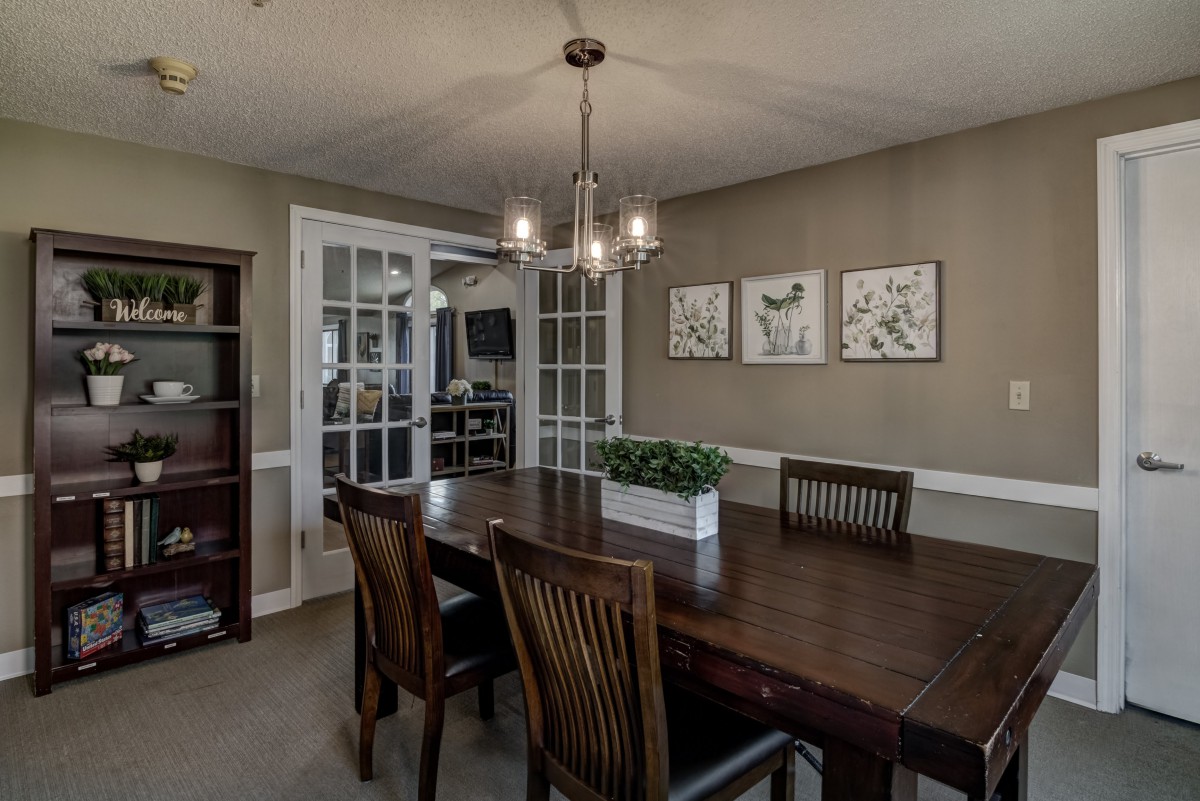A modern dining room featuring a wooden table and chairs, illuminated by a stylish chandelier, with artwork and plants enhancing the decor.