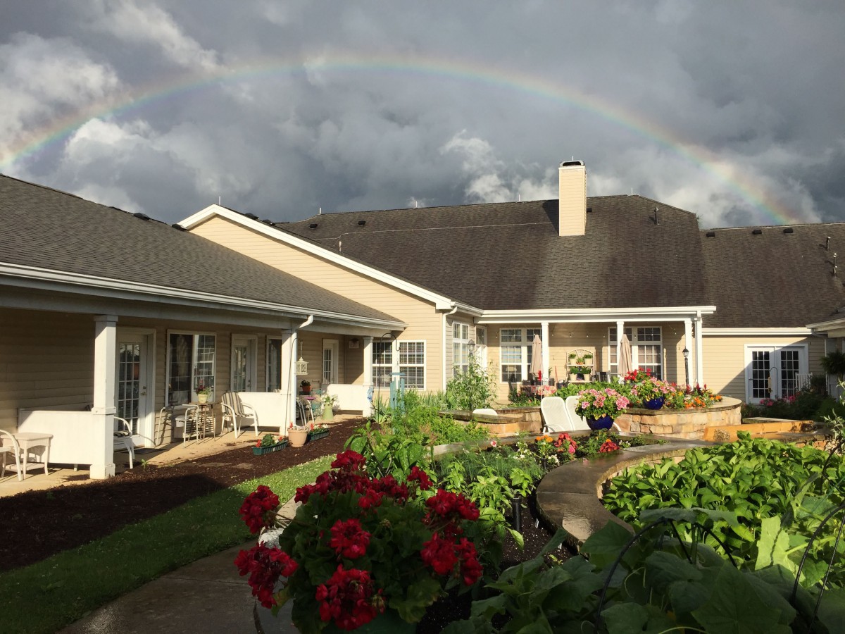 A peaceful garden view with a rainbow arching over houses and vibrant flowers.