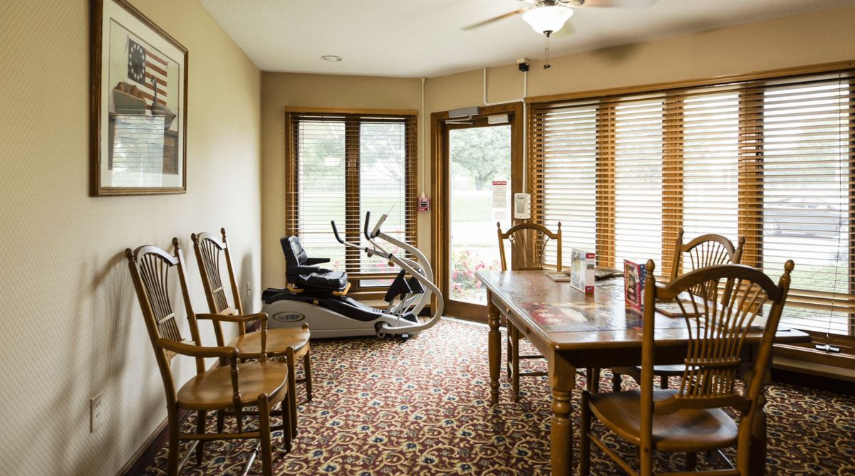 A room with an exercise bike next to a table and chairs, featuring large windows and a ceiling fan.