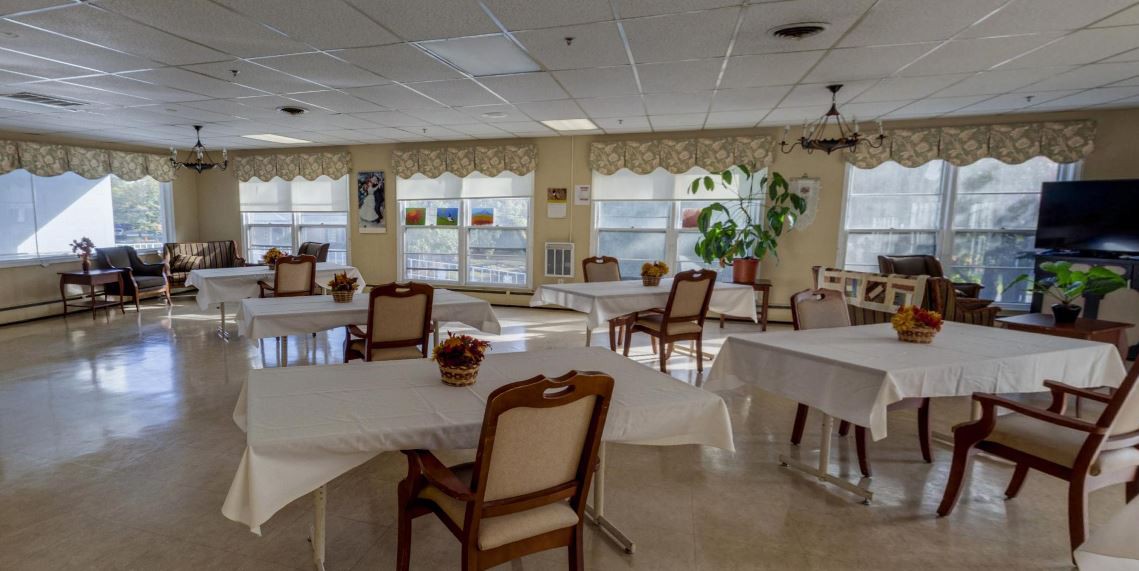 An empty dining room with round tables, chairs, and large windows letting in natural light.
