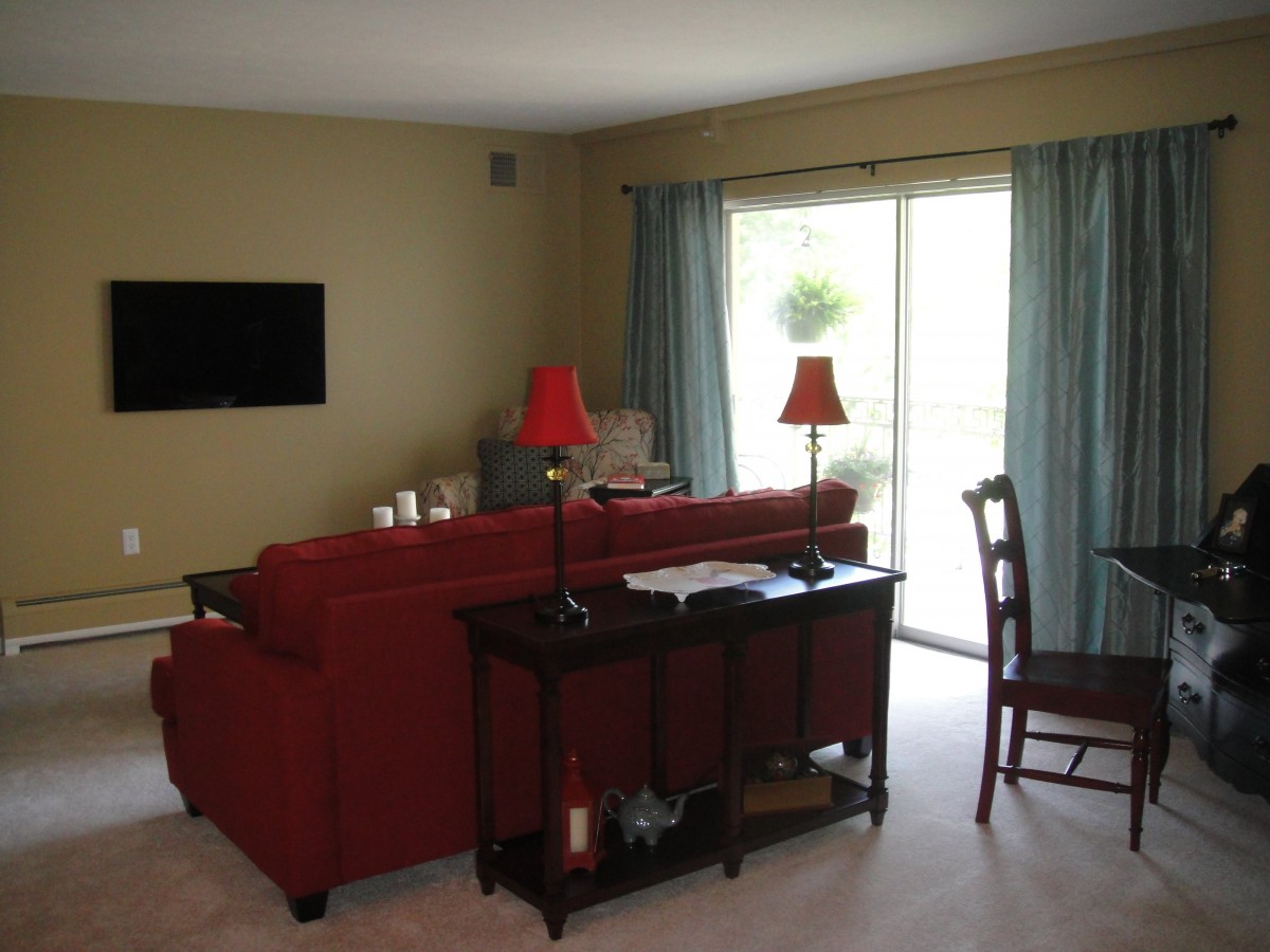 A cozy living room with a red sofa, lamps, and a television mounted on the wall. The room has a sliding glass door with blue curtains.