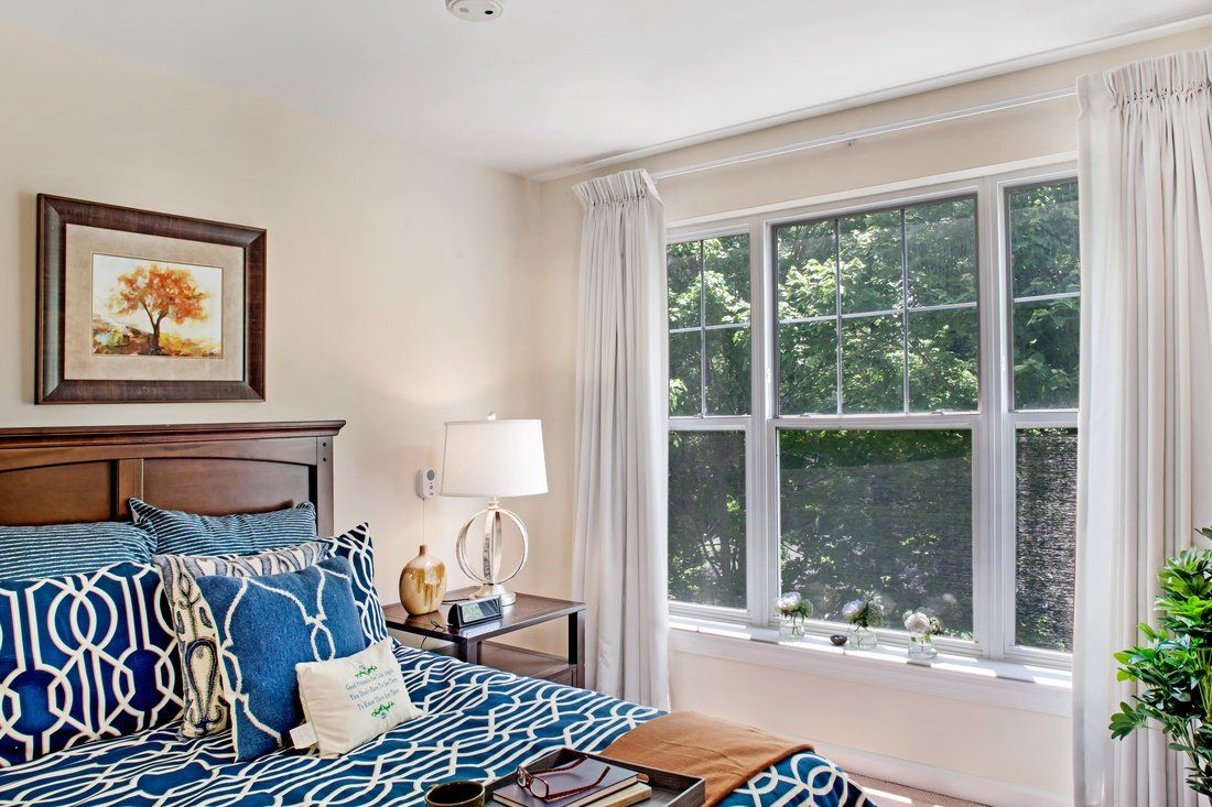 A cozy bedroom with a large window, decorated in blue and white tones, featuring a bed with patterned bedding and a bedside lamp.