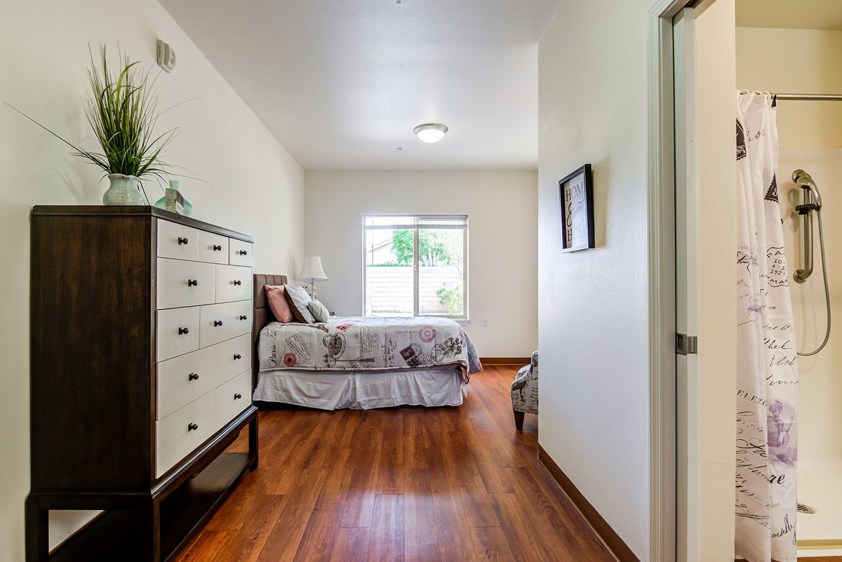 A cozy bedroom with a bed, dresser, and hardwood flooring, featuring natural light from a window.