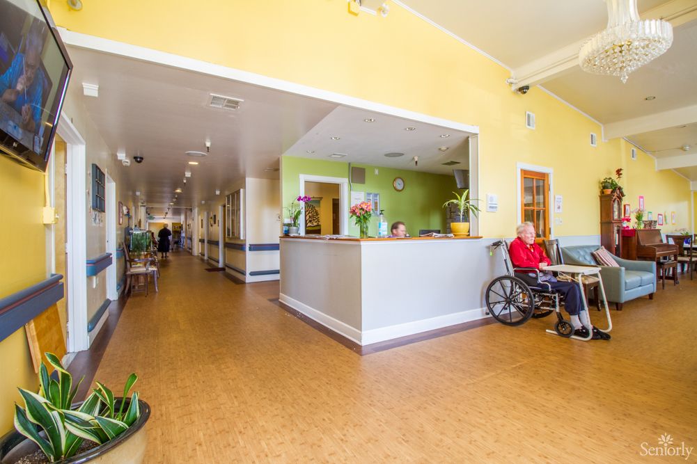 A bright and welcoming nursing home reception area with a person in a wheelchair and a view down the hallway.