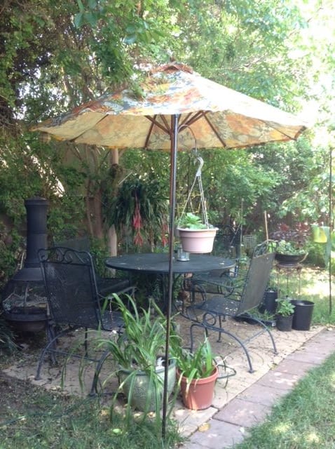 A cozy garden patio with a table, chairs, umbrella, and chiminea surrounded by lush greenery.