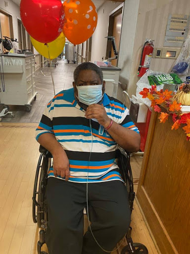 A person in a wheelchair wearing a mask, seated indoors with festive balloons in the background.