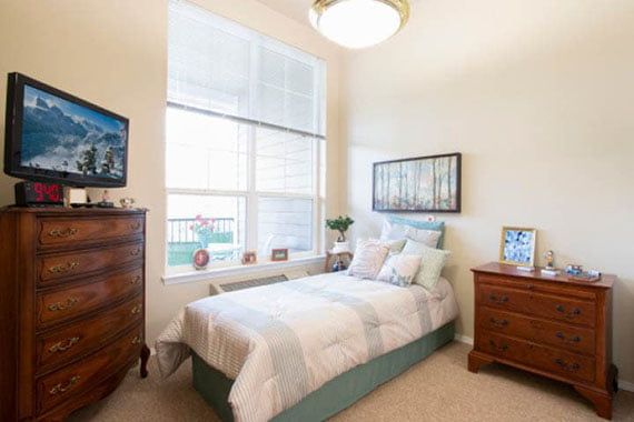 A cozy bedroom with a large window, featuring a single bed, wooden furniture, and soft lighting.