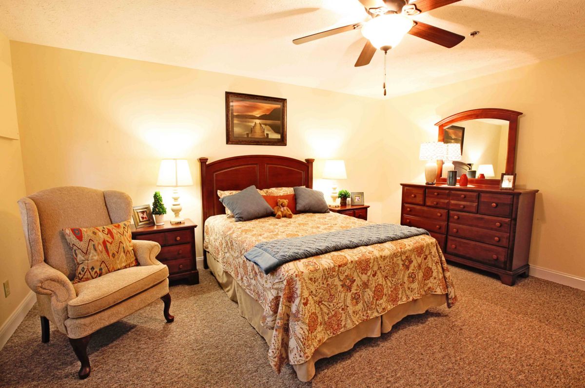 A cozy bedroom with warm lighting, featuring a bed, armchair, dresser, and decorative elements.