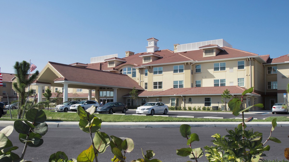 A mid-sized hotel with a brown roof, featuring parked cars and surrounding greenery under a clear blue sky.