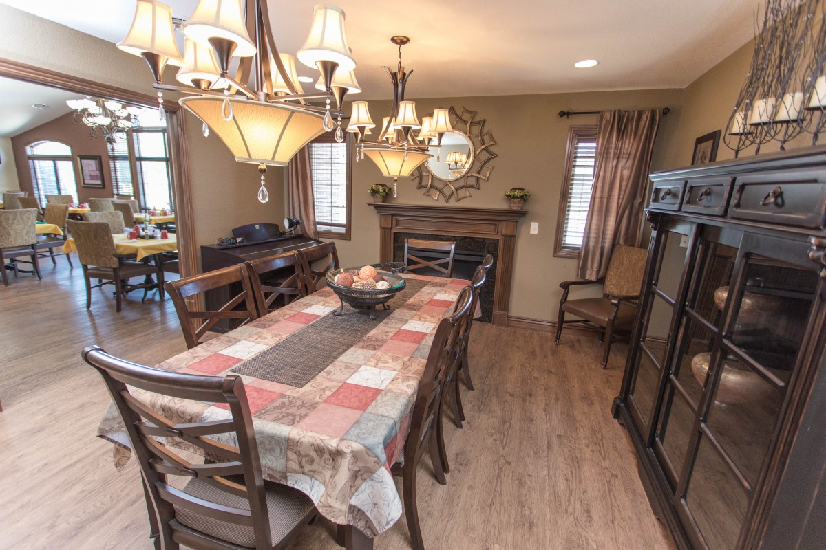 A cozy dining room featuring a long table with a colorful tablecloth, surrounded by chairs. A chandelier hangs overhead, and a fireplace with a mirror above it adds warmth to the space.