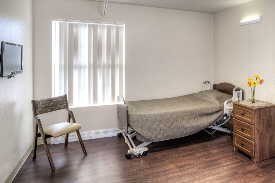 A minimalist room with a hospital bed and chair, featuring a wooden floor and window with vertical blinds.