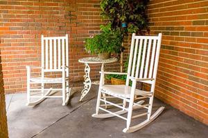 Two white rocking chairs and a small table adorn a cozy patio area against a brick wall, accented by lush greenery.