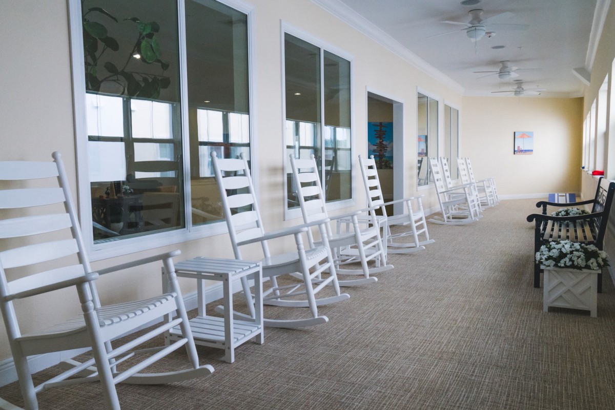 A row of white rocking chairs lined up along a porch with large windows.