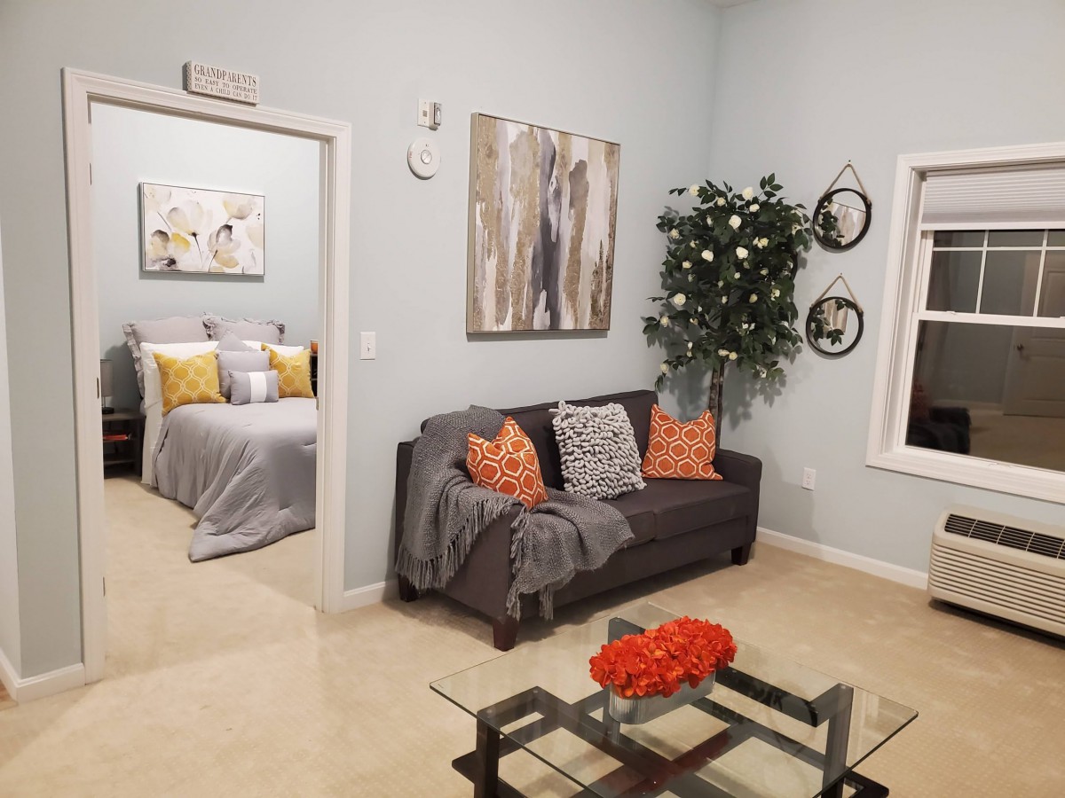 A cozy living room with a gray sofa, colorful pillows, and modern artwork. A glimpse into the bedroom is visible through an open doorway.