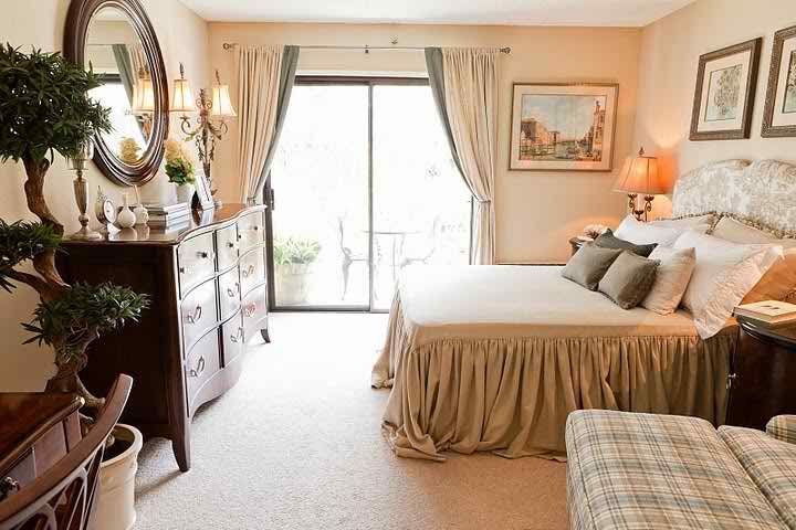 A cozy and elegantly decorated bedroom featuring a large bed, wooden dresser, and soft carpeting. Natural light filters through the sliding glass door adorned with curtains.