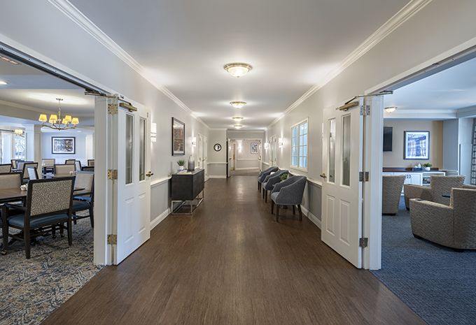 A well-lit hallway with wooden flooring and multiple doors, featuring chairs arranged along the side.
