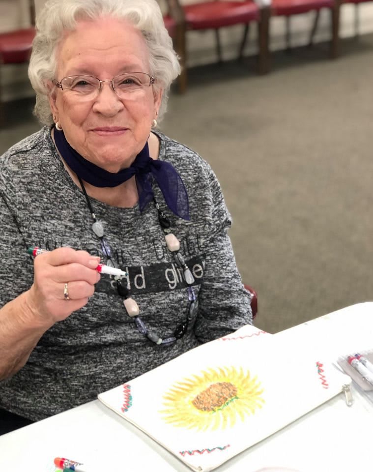 A smiling elderly woman engaged in an art activity, holding a marker and working on a colorful drawing.