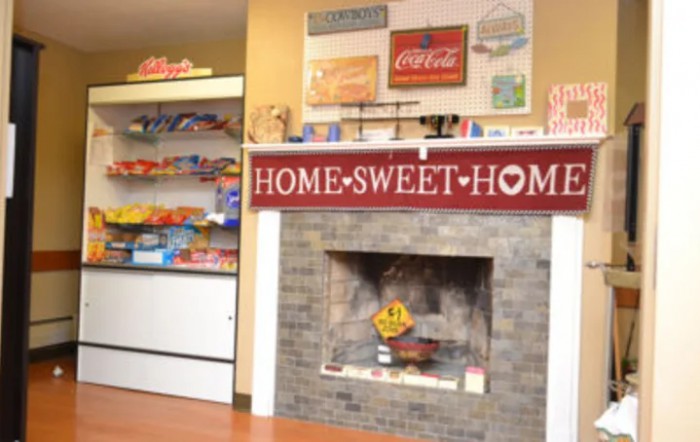 A cozy interior featuring a 'Home Sweet Home' sign above a fireplace, with shelves of food items and vintage Coca-Cola signage.