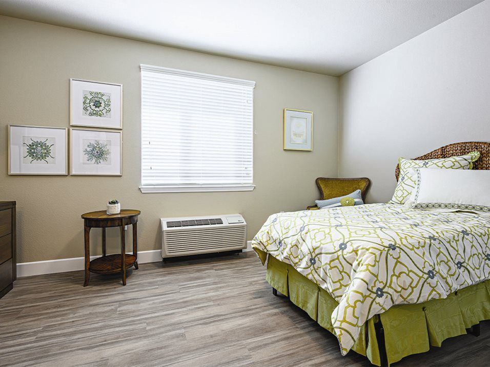 A cozy bedroom with a neatly made bed, artwork on the walls, and a wooden side table by the window.