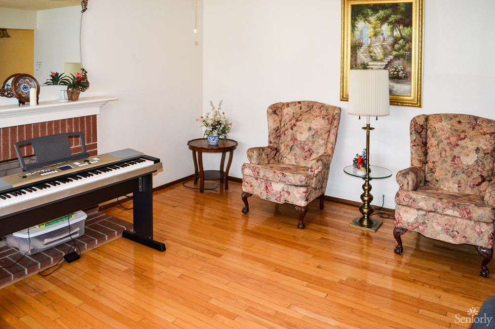 A cozy living room with a digital piano, two floral armchairs, a lamp, and a painting on the wall.