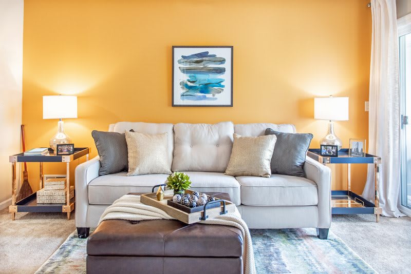 A cozy living room with a light gray sofa, accented by colorful cushions, set against a vibrant yellow wall featuring modern artwork. Two side lamps provide warm lighting, complementing the stylish decor.