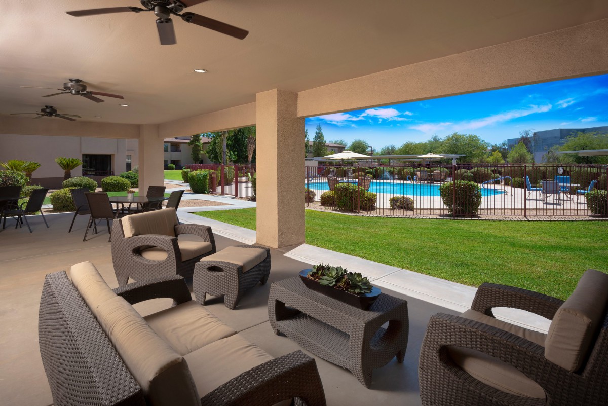 A covered patio with comfortable seating overlooks a pool and landscaped garden.