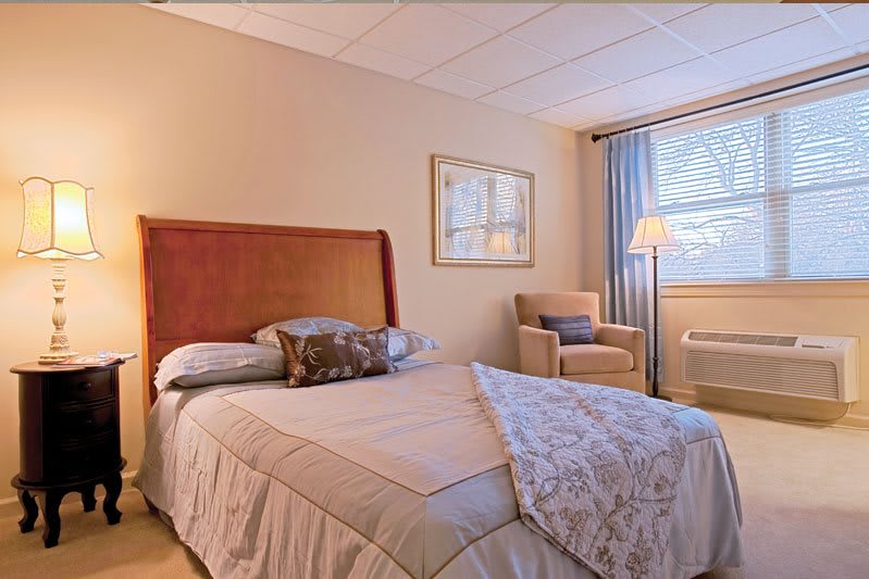 A cozy bedroom with a neatly made bed, an armchair by the window, and soft lighting from a floor lamp.