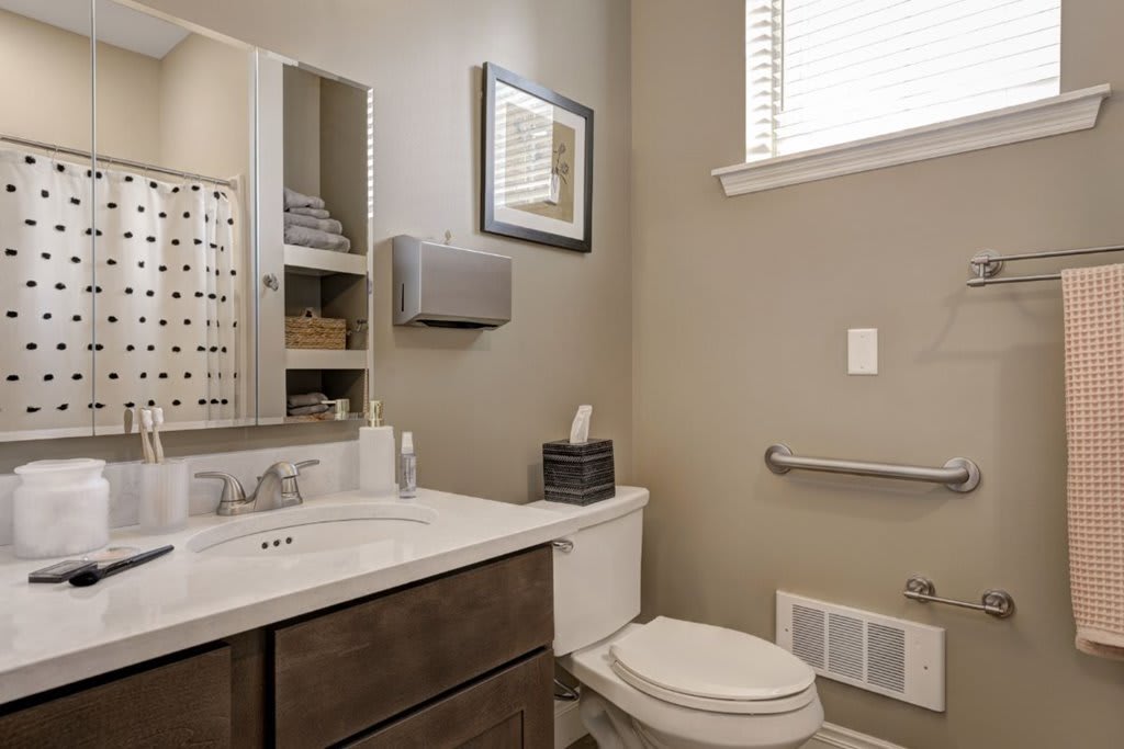 A modern bathroom with a toilet, sink, and mirror, featuring beige walls and an accessible handrail.