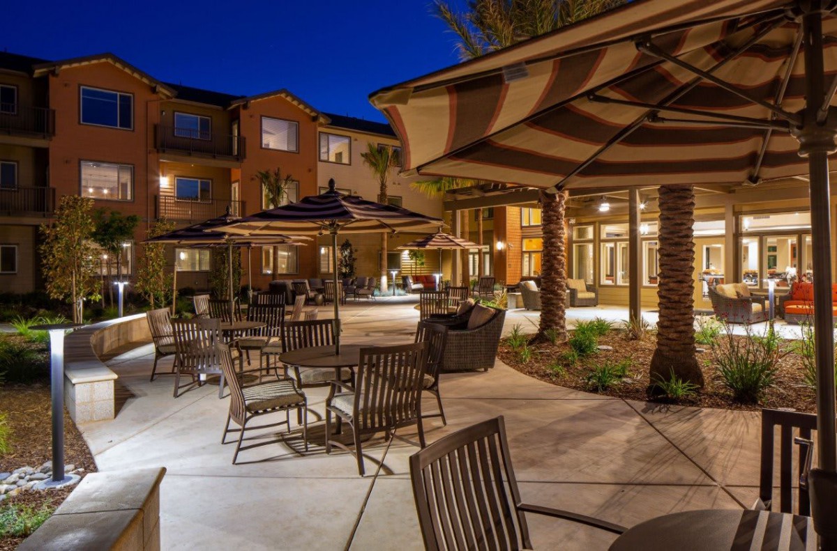 A well-lit outdoor patio with tables and chairs set under umbrellas by a building in the evening.