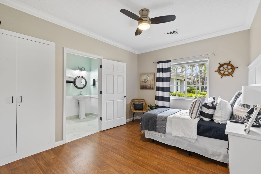 A modern bedroom with a ceiling fan, wooden flooring, and abundant natural light coming through a window with striped curtains.