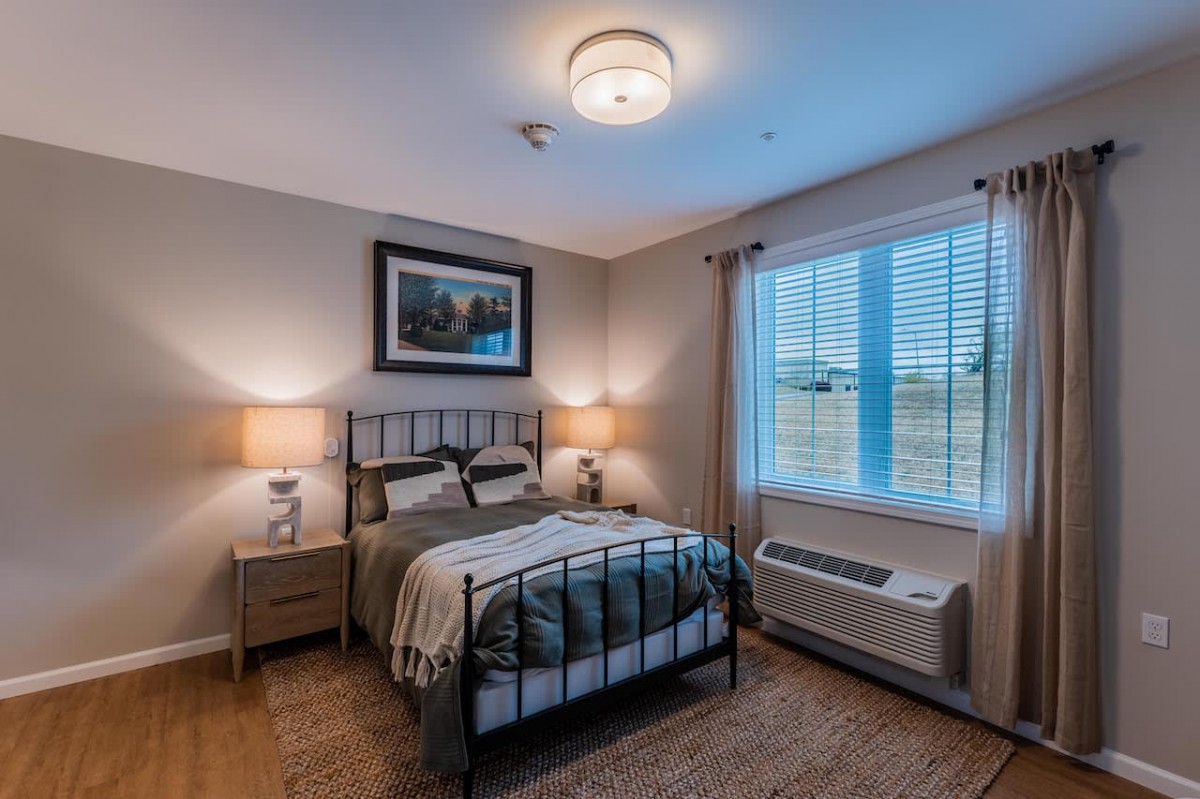 A cozy and modern bedroom with warm lighting, featuring a comfortable bed, two bedside lamps, and a window with blinds.