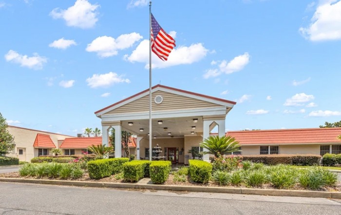 A building entrance with a flagpole displaying an American flag, surrounded by neatly arranged plants and shrubs.