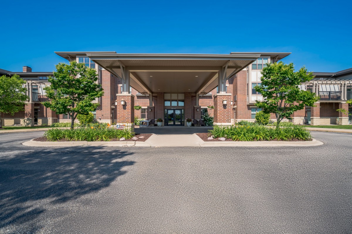 The image shows the front entrance of a modern building with a covered driveway, surrounded by well-maintained landscaping and trees under a clear blue sky.