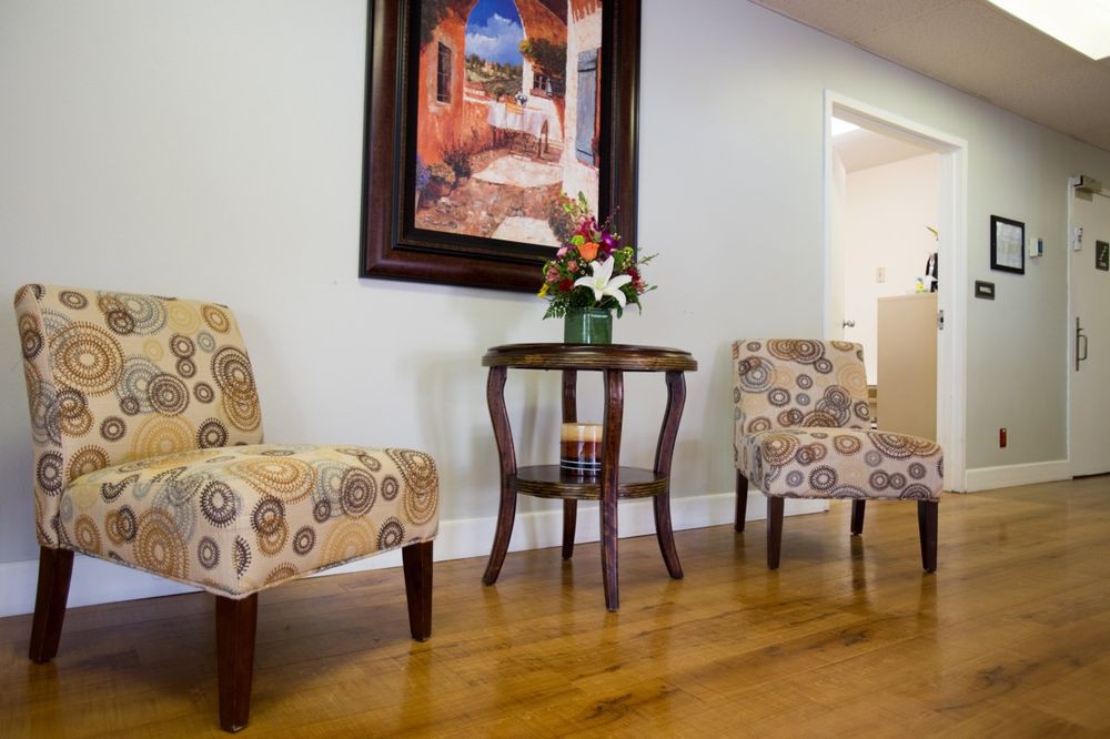 A cozy waiting area with patterned chairs and a small table adorned with flowers beneath a colorful painting.