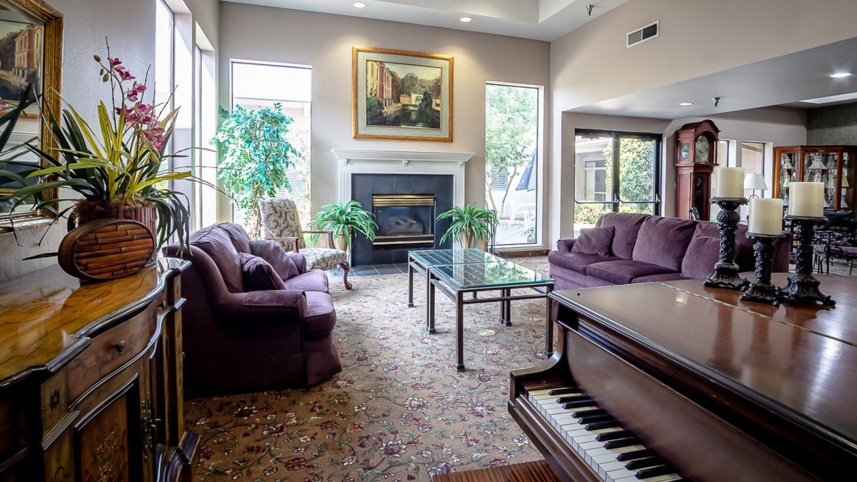 A cozy living room with plush sofas, a piano, and a fireplace, decorated with plants and artwork.