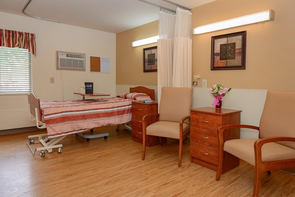 A clean and well-organized hospital room with a single bed, chairs, and a small table with flowers.