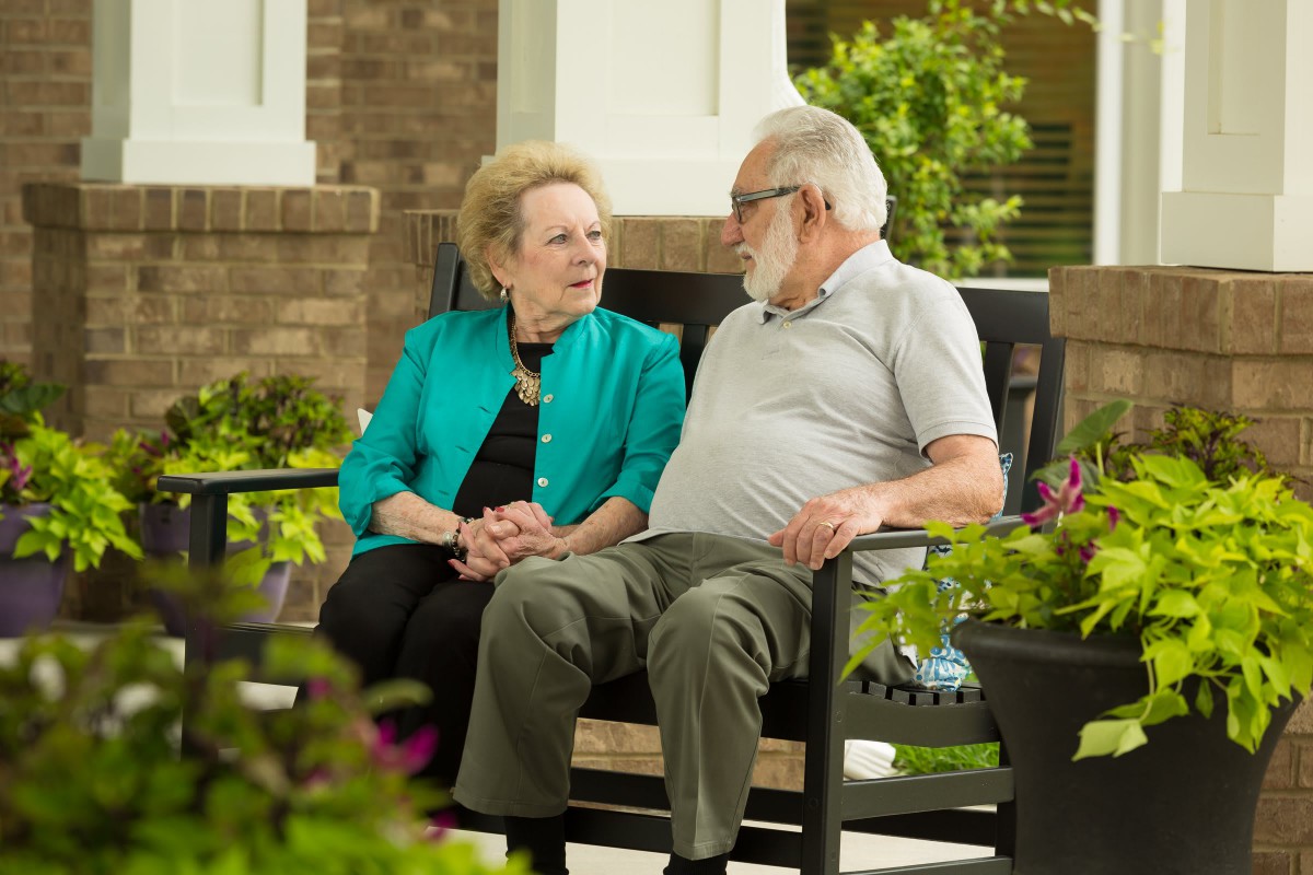 An elderly couple sitting on a bench having a conversation in a garden setting.