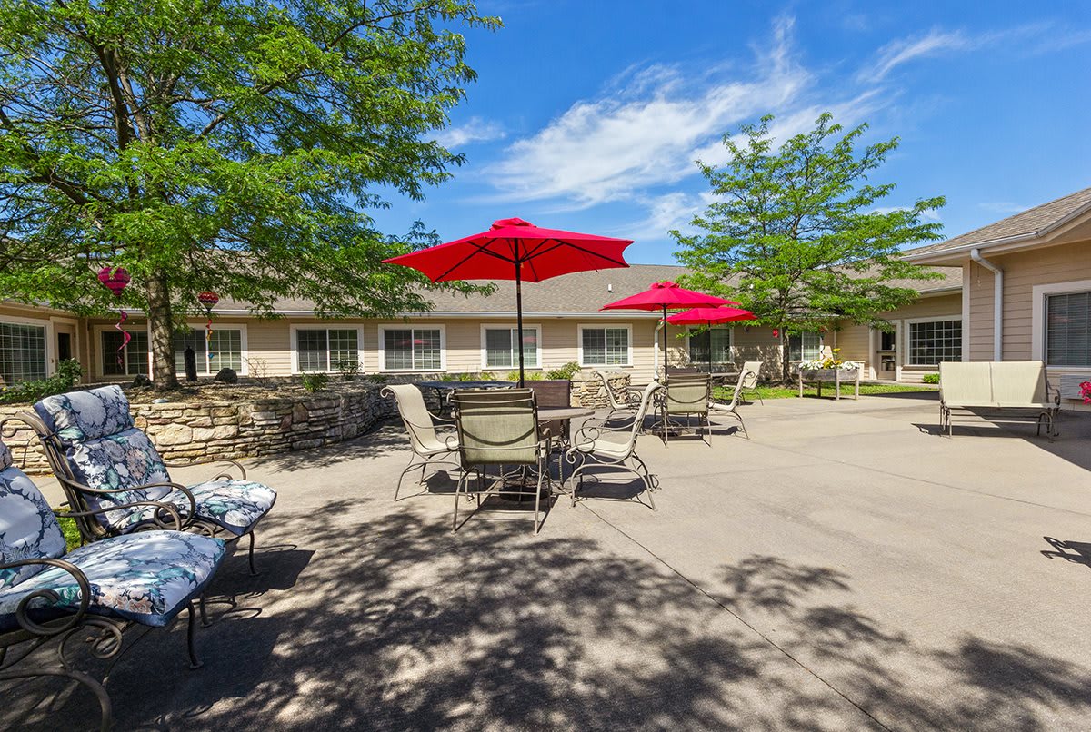 A sunny outdoor patio area with tables, chairs, and red umbrellas near a building.