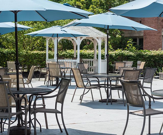 A peaceful patio area with tables, chairs, and umbrellas set up under a bright sun, complemented by a white gazebo and lush greenery in the background.