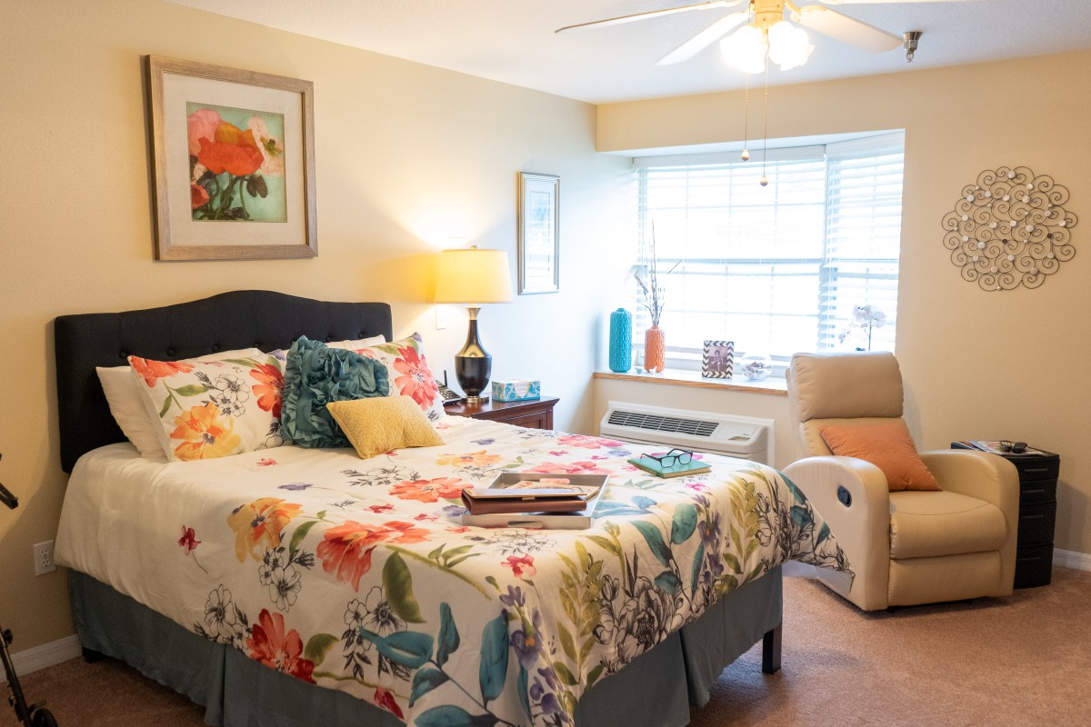 A cozy bedroom with colorful floral bedding, an armchair by the window, and warm lighting.