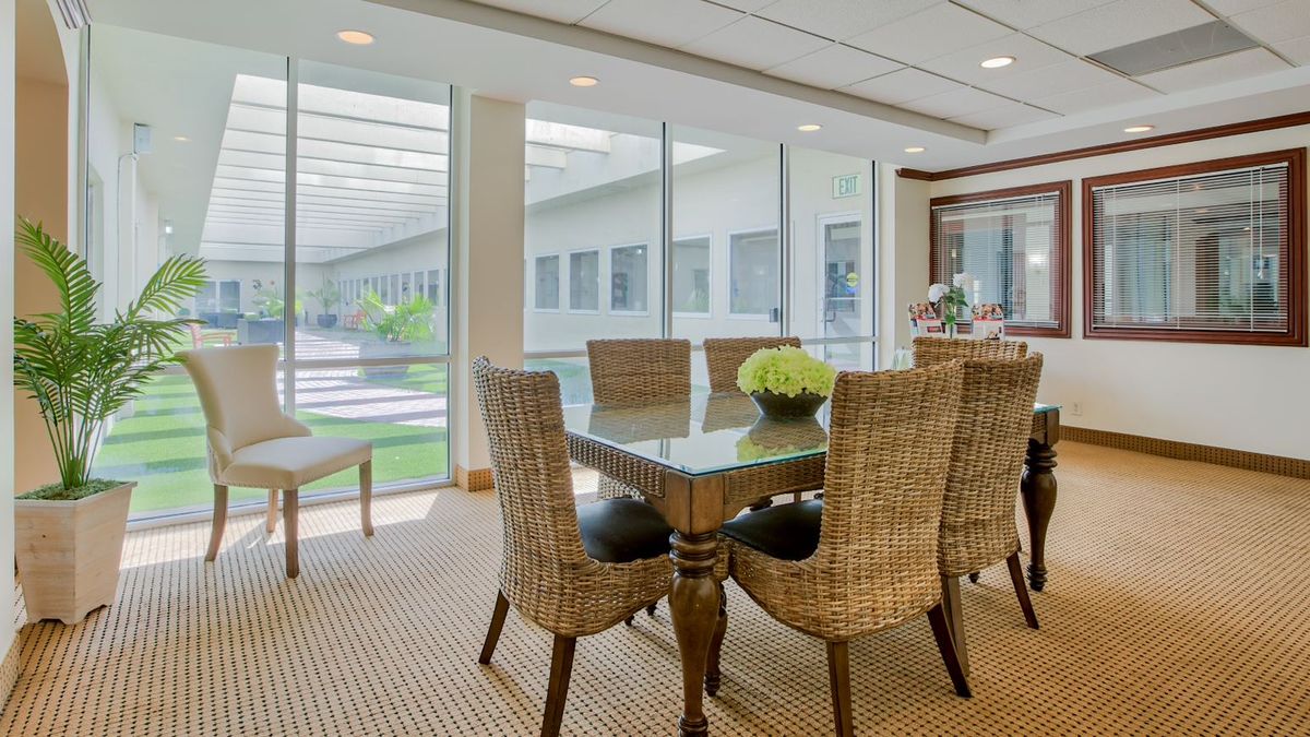 A modern dining room with wicker chairs and a glass table, well-lit by large windows overlooking a garden.