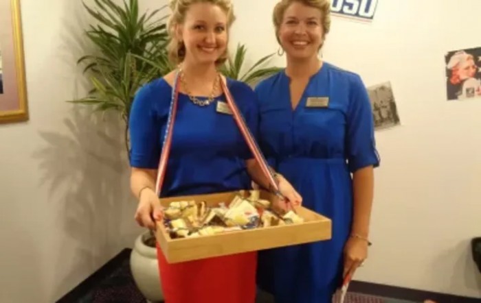 Two women wearing uniforms, one holding a serving tray with food items, smiling at an event.