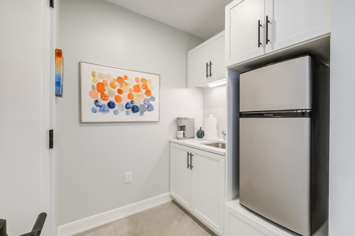 A modern kitchen area with white cabinets, a refrigerator, and colorful artwork on the wall.