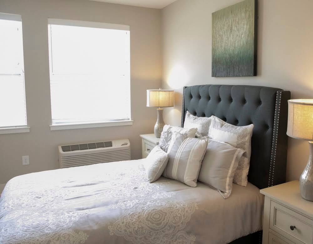 A well-decorated bedroom with a plush bed, ample pillows, a bedside table with a lamp, and a window allowing natural light.