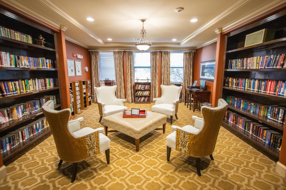 A cozy library reading room with comfortable chairs, a central table, and shelves filled with books.