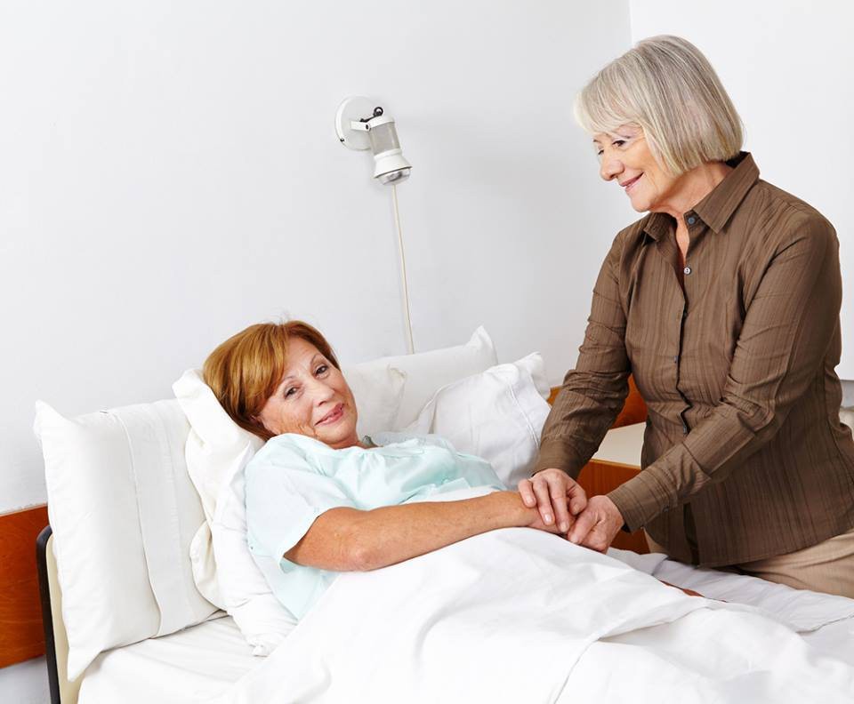 A supportive visitor holding hands with a patient lying in a hospital bed.