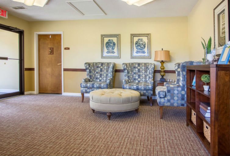 A cozy waiting room area featuring patterned chairs, a round ottoman, and framed artwork on the wall.